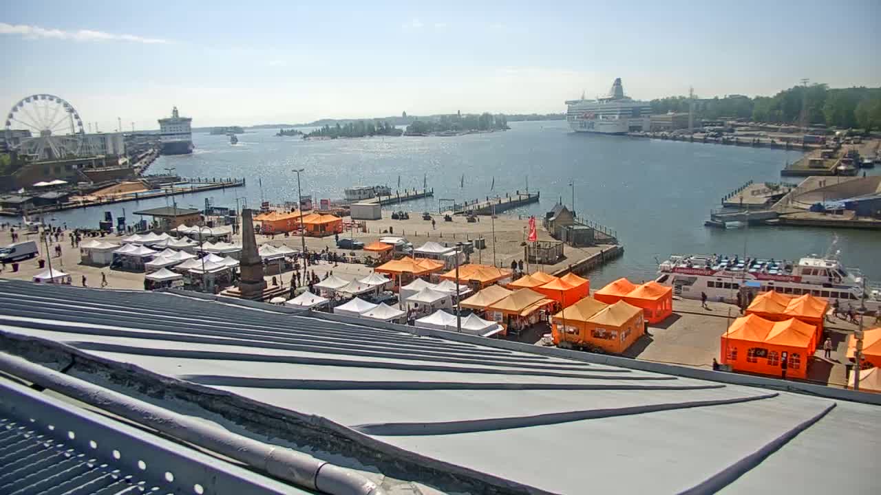 A harbor scene on a sunny day features numerous orange and white market tents situated near a dock, with a ferry and a Ferris wheel visible in the distance.