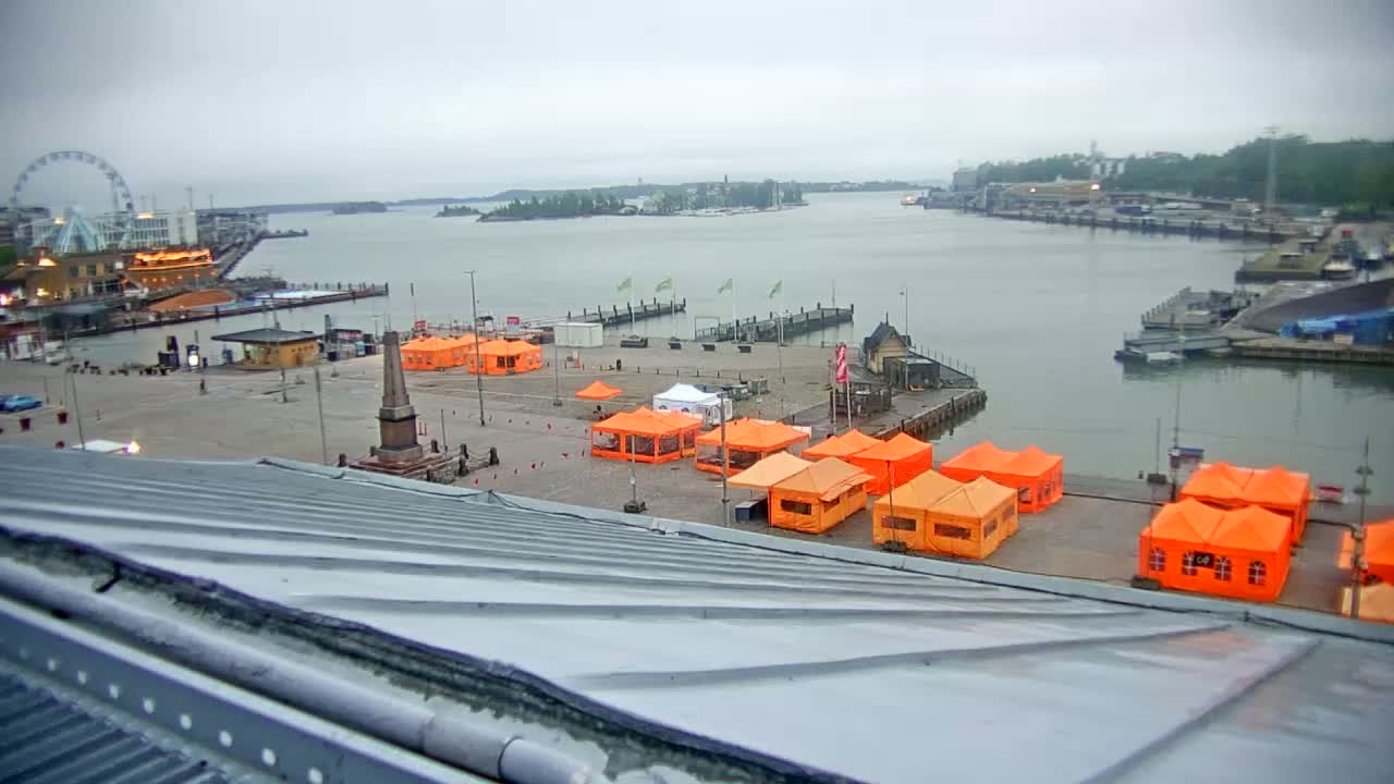 A harbor scene on an overcast day shows numerous orange tents set up on a paved area next to a body of water, with a Ferris wheel and buildings visible in the background.