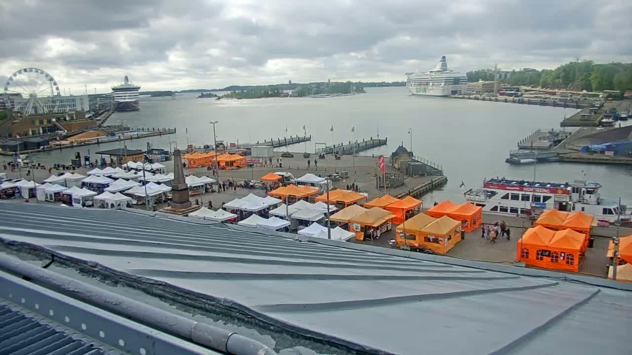An outdoor market with orange and white tents is situated near a harbor with docked cruise ships and a Ferris wheel, under a cloudy sky.