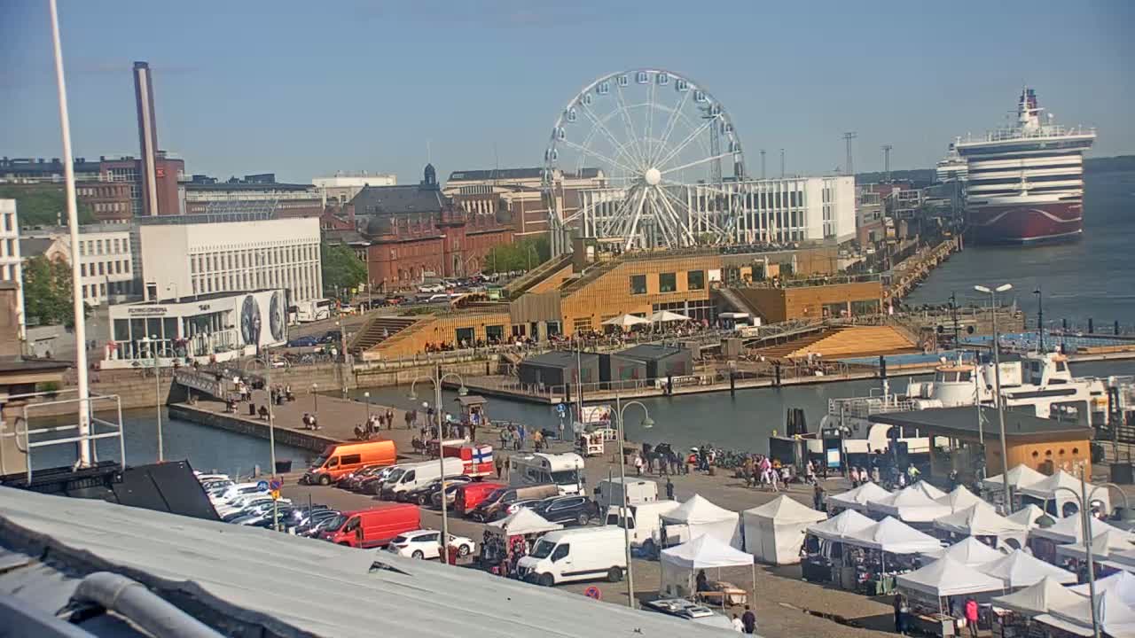A waterfront scene on a sunny day shows a Ferris wheel, a cruise ship, numerous market stalls, parked cars, and people strolling along a harbor.
