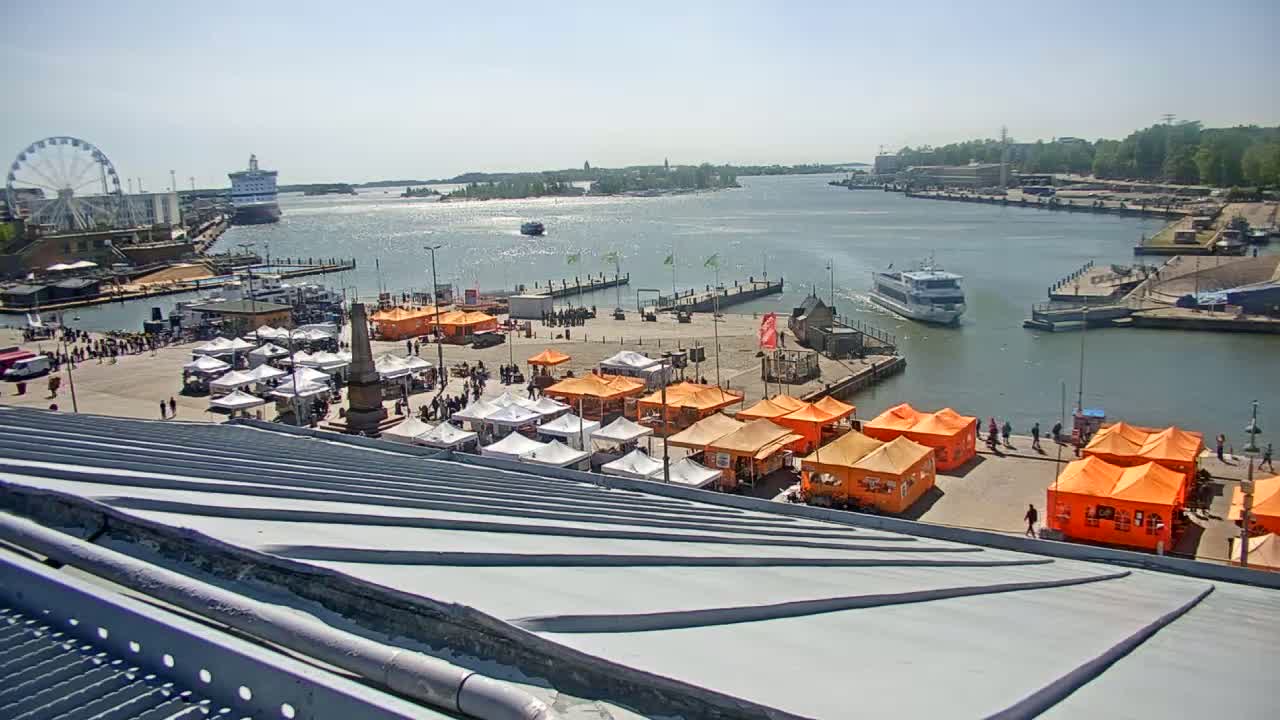 A harbor scene on a sunny day shows numerous orange and white market stalls surrounding a harbor with boats and a Ferris wheel in the background.