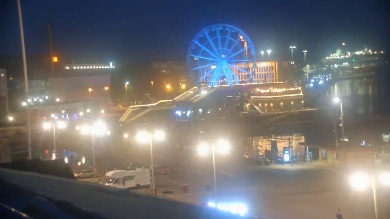 A brightly lit waterfront at night features a large Ferris wheel illuminated in blue, with numerous streetlights and parked vehicles visible in the foreground.