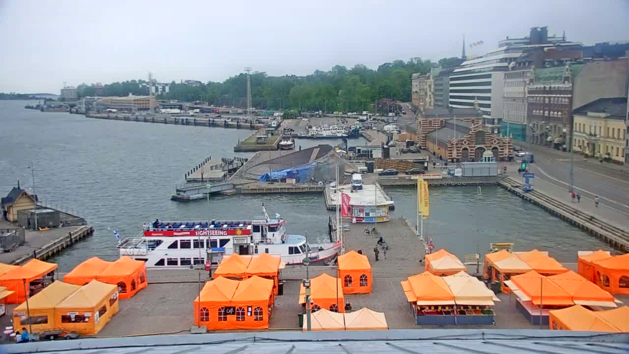 An overcast day reveals a harbor scene with several docked boats, orange market tents, and buildings lining a waterfront street.