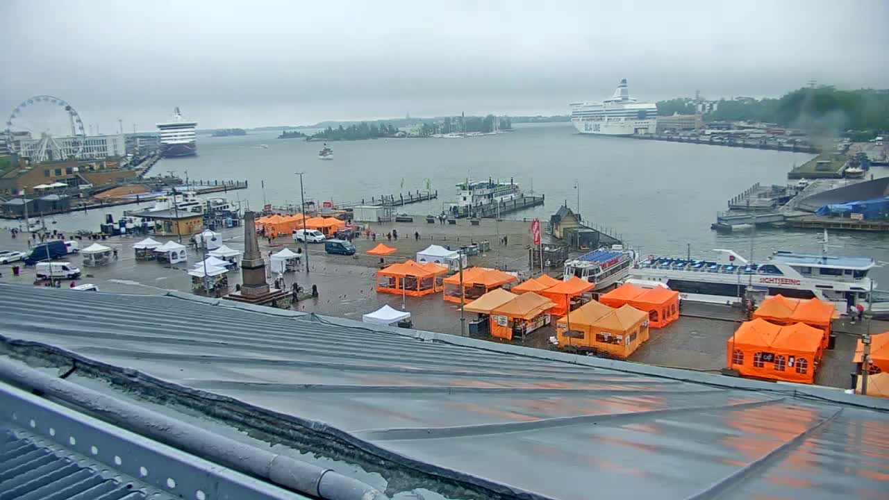 A harbor scene on an overcast day shows numerous orange market stalls in a square, boats and ships in the water, and a Ferris wheel in the distance.