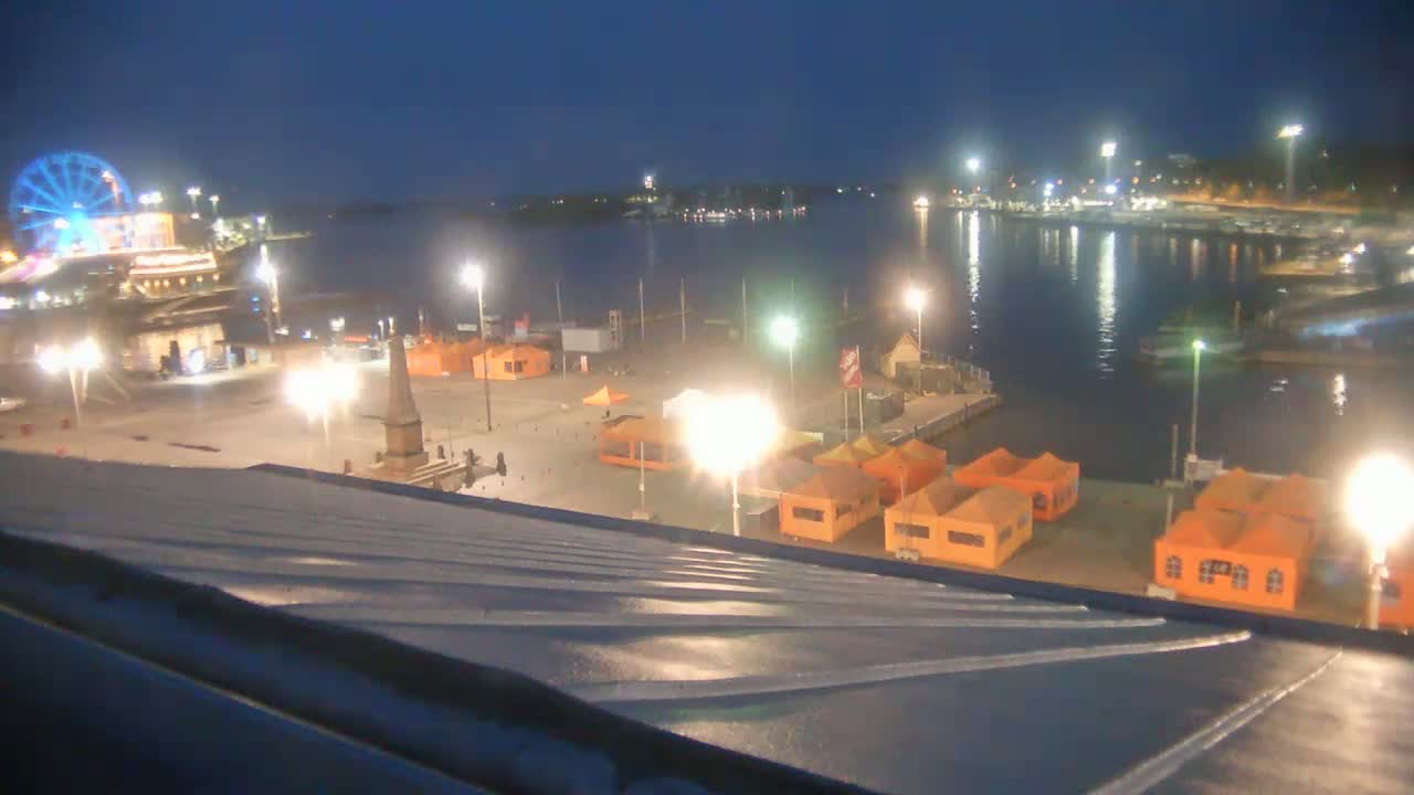 A nighttime view from a rooftop shows a brightly lit waterfront area with a Ferris wheel, numerous orange tents, and a calm body of water.
