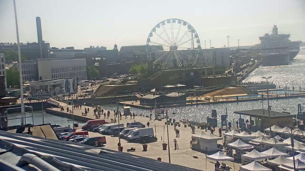 A sunny day at a harbor shows a Ferris wheel, a docked large ship, several buildings, a paved area with people and cars parked, and numerous market stalls.