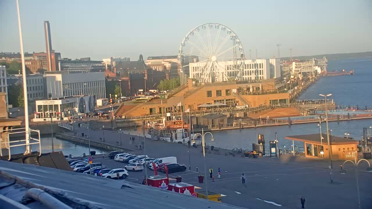 A waterfront area with a Ferris wheel, buildings, a boat, cars in a parking lot, and calm water under a clear, light sky.