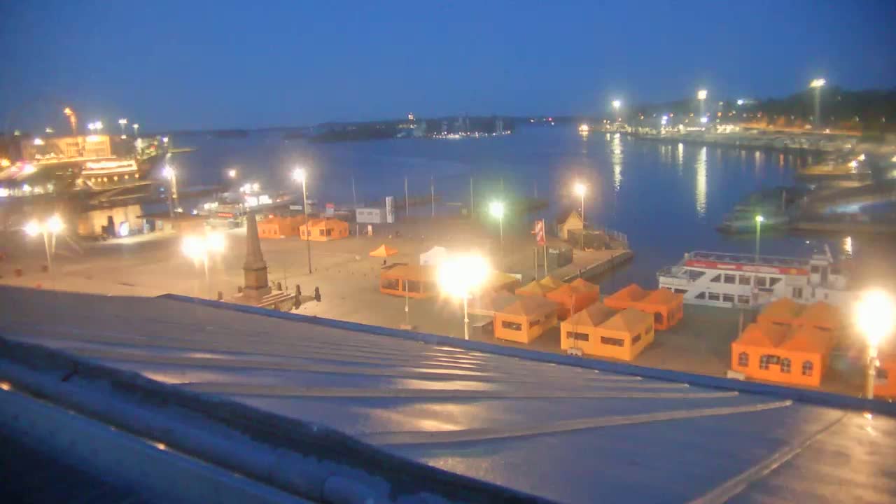 A nighttime view of a harbor with boats docked, orange tents, and lights illuminating a plaza, overlooking a calm body of water under a twilight sky.