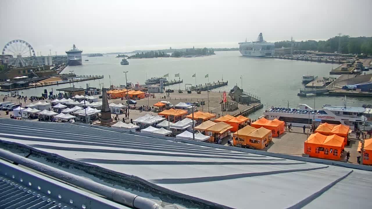 A harbor scene on a hazy day shows numerous orange and white market stalls, a Ferris wheel, several ships, and a sightseeing boat.