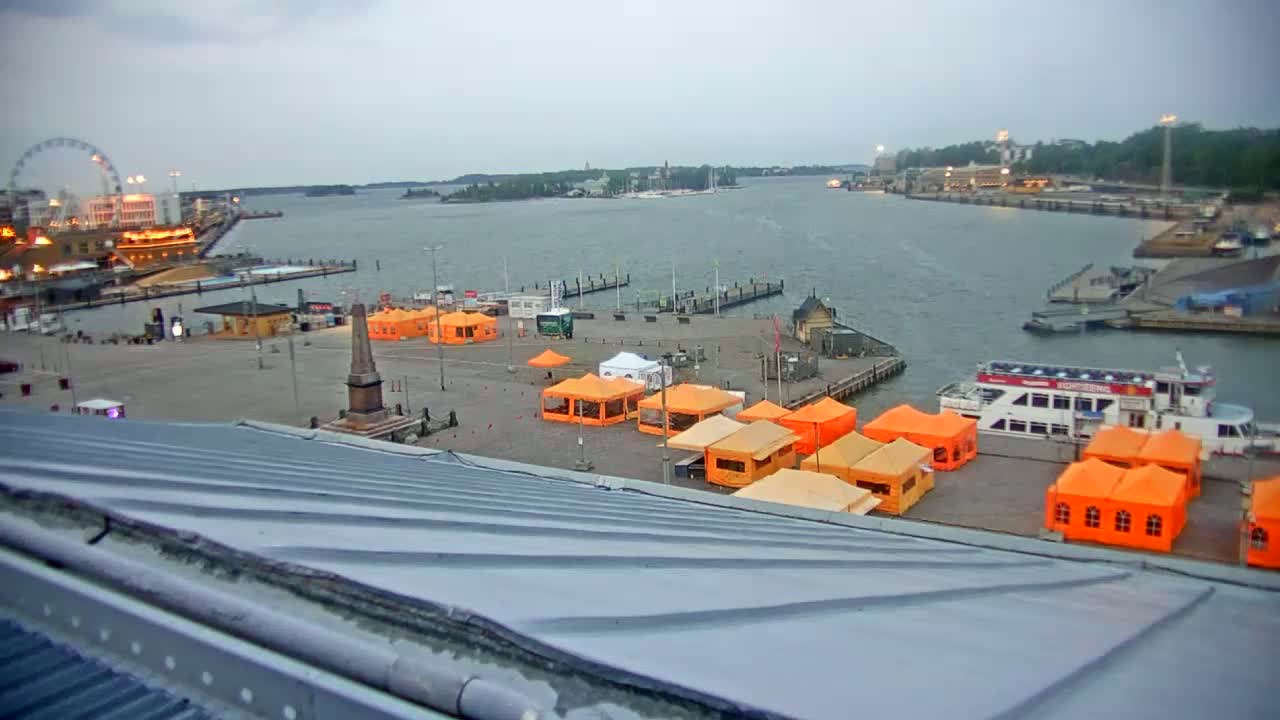 A harbor scene with numerous orange tents set up on a plaza, a ferry docked, a Ferris wheel in the distance, and overcast skies.
