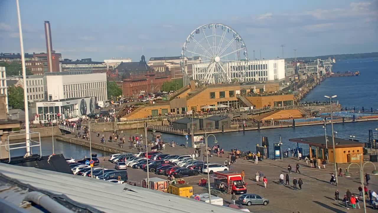 A waterfront area with a Ferris wheel, buildings, a parking lot filled with cars, and many people walking around on a sunny day.