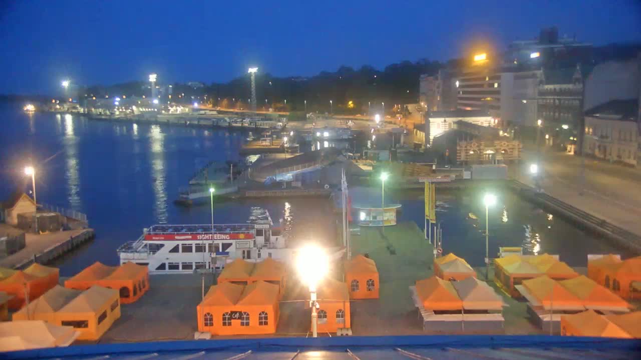 A nighttime view of a harbor filled with docked boats and illuminated orange tents, under a dark blue sky.