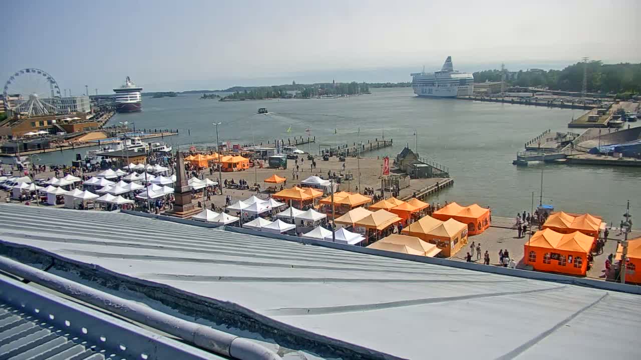 A harbor scene on a sunny day shows numerous market stalls set up on a waterfront plaza, with ferries and a Ferris wheel in the background.
