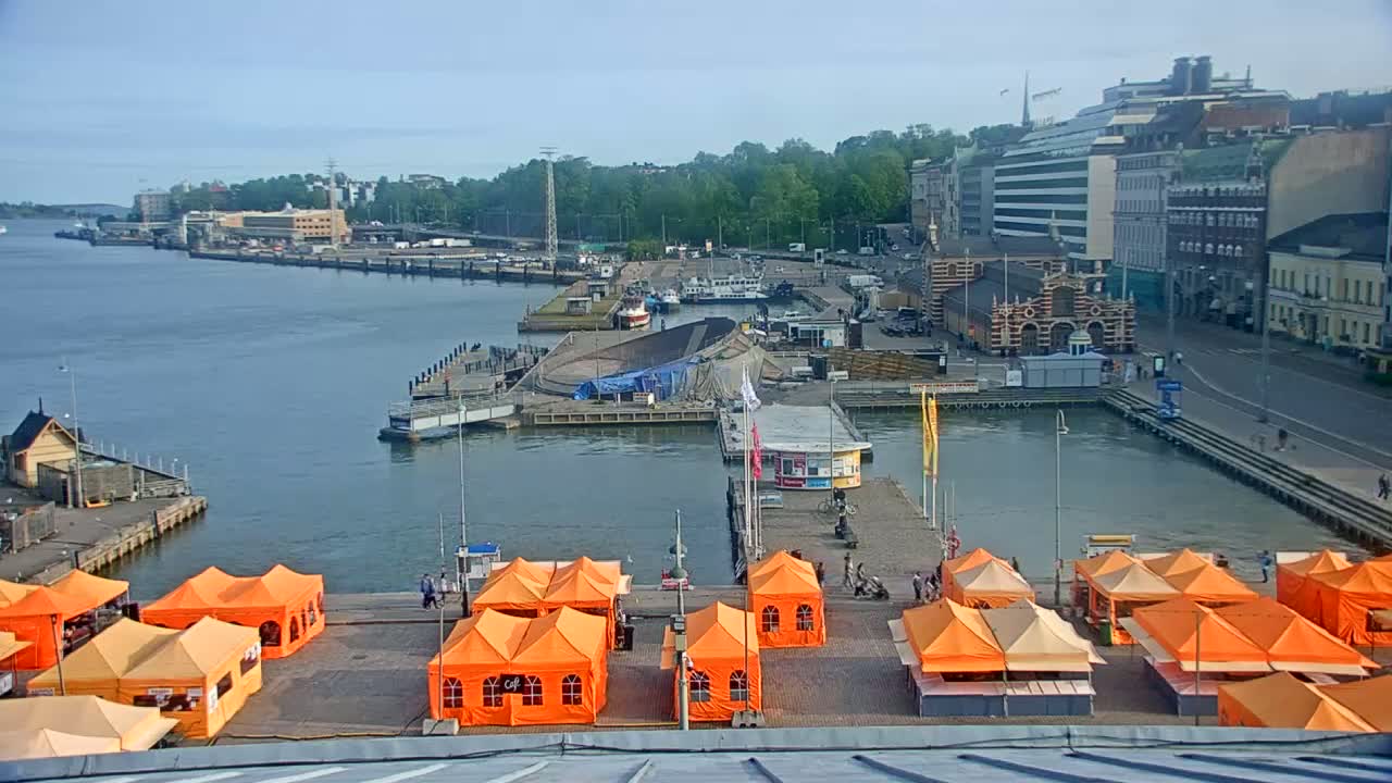 A harbor scene on a clear, sunny day shows numerous orange market tents situated on a pier, with boats and buildings in the background.