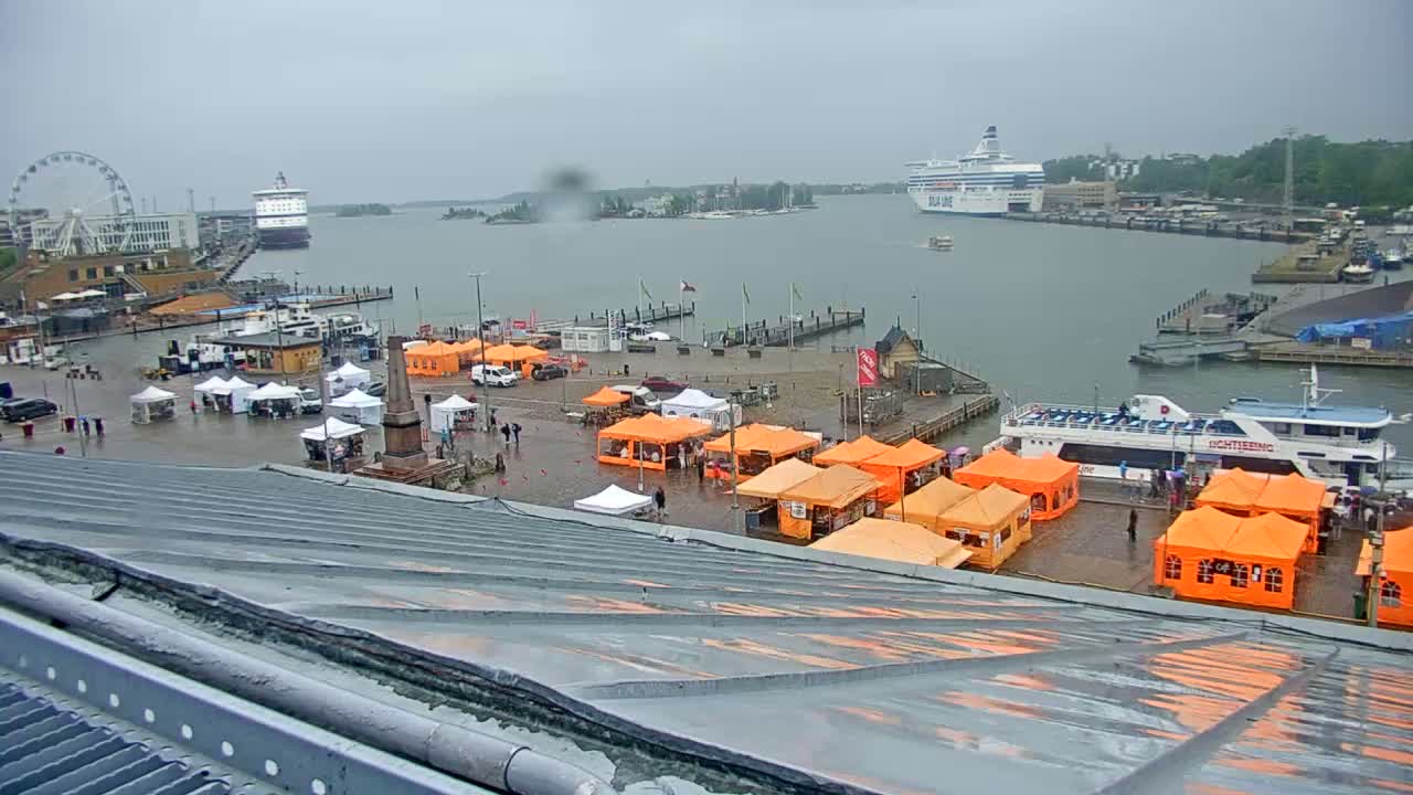 A harbor scene on an overcast day shows numerous orange tents set up in a paved area near a waterfront with several ships, including a large ferry, and a Ferris wheel in the background.