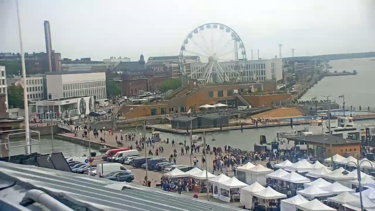 A Ferris wheel overlooks a waterfront area with numerous people, market stalls, and boats on an overcast day.