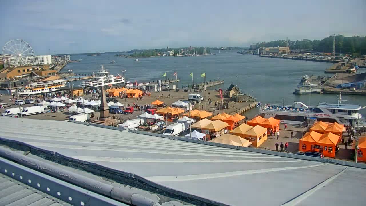 A waterfront market with numerous orange tents is situated near docked ferries and boats on a sunny day.