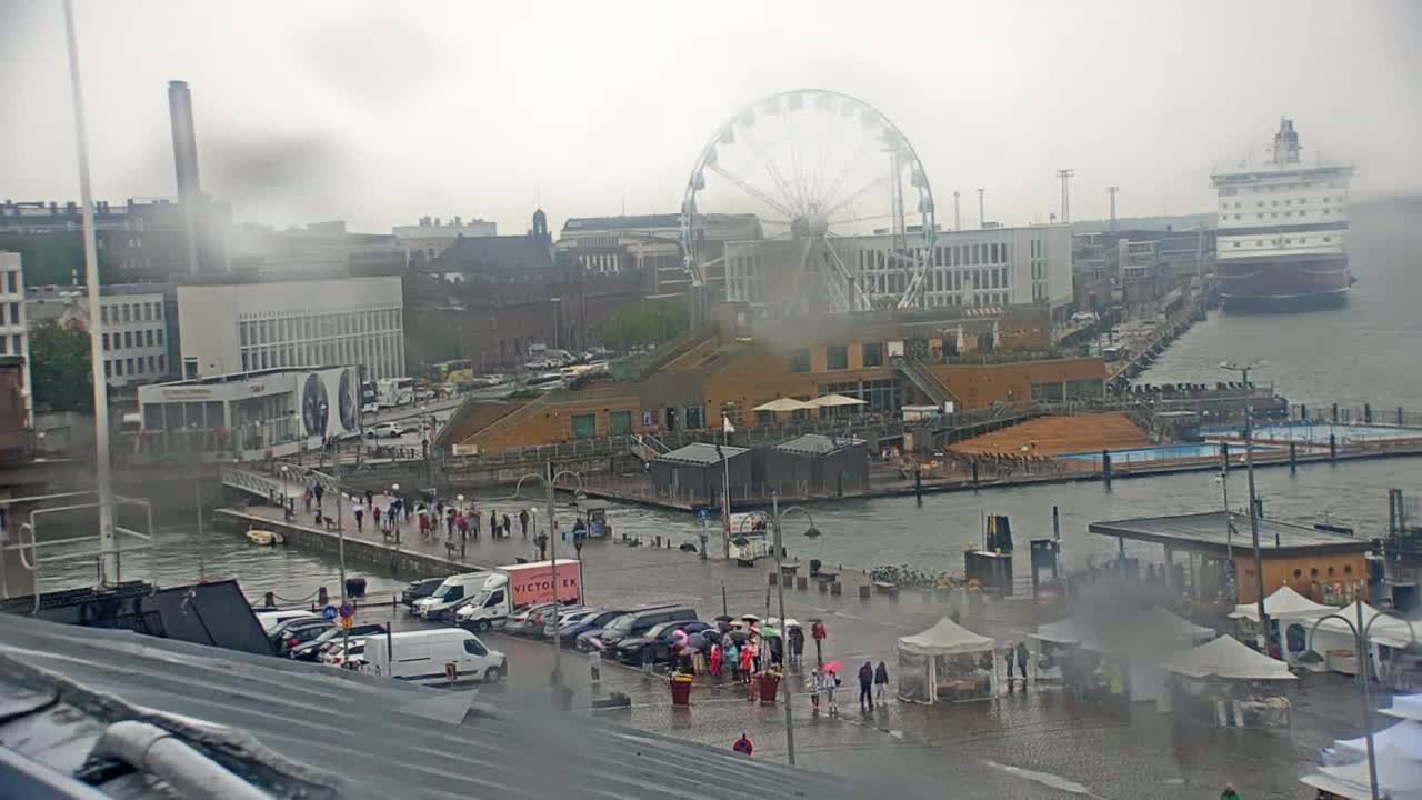 A waterfront city scene on a rainy day shows a Ferris wheel, a cruise ship, buildings, people walking along a promenade, and various small structures and market stalls near a body of water.