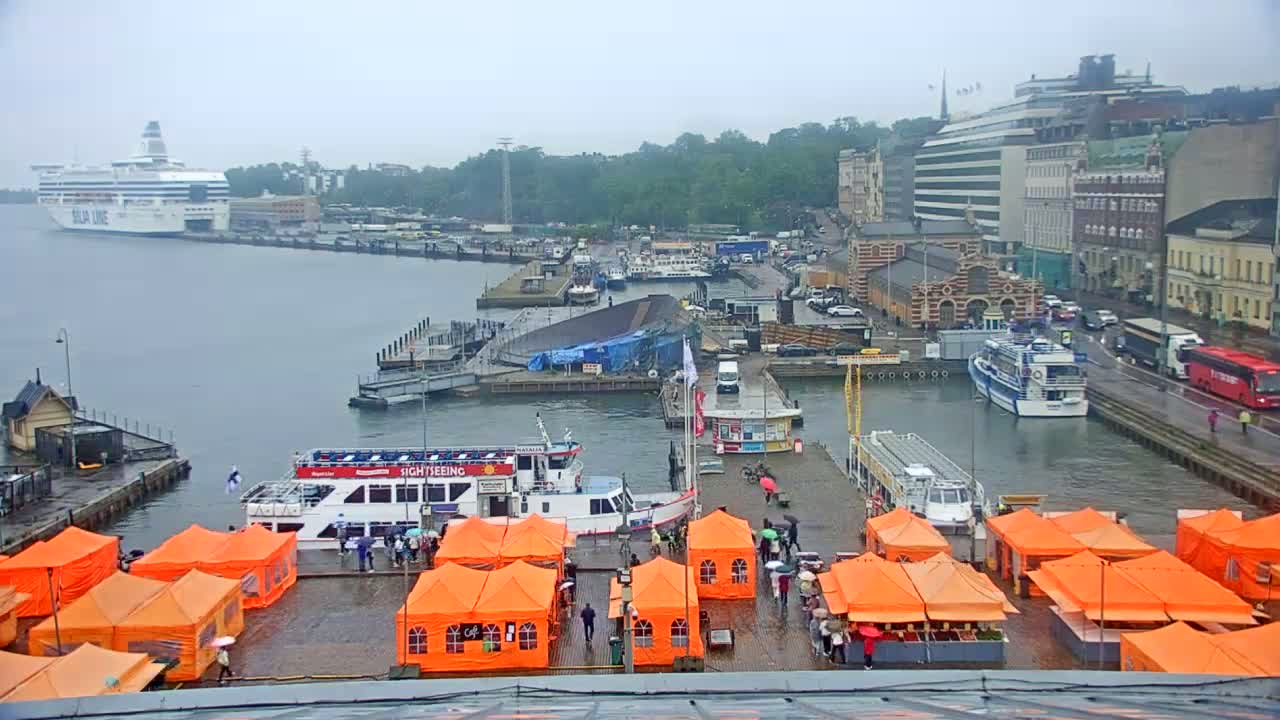 A harbor scene on a rainy day shows several boats and a large cruise ship docked near buildings and a market with orange tents.
