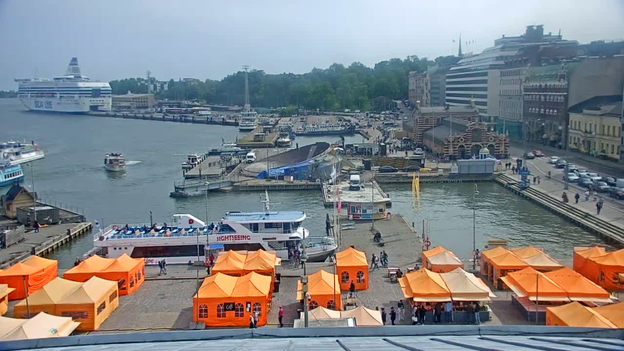 A harbor scene on an overcast day shows several boats, including a large cruise ship, docked near buildings and orange market tents.
