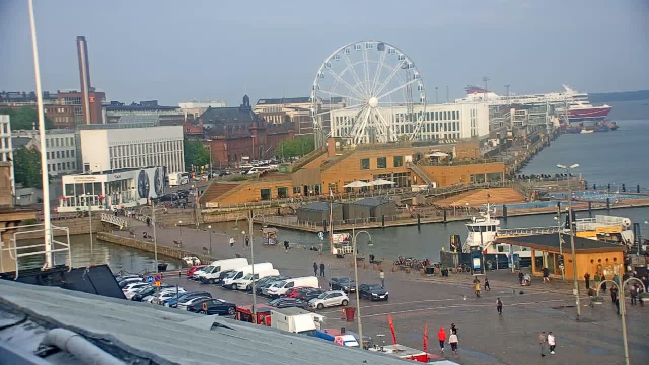 An overcast day at a waterfront harbor with a Ferris wheel, buildings, docked boats, and people walking around.