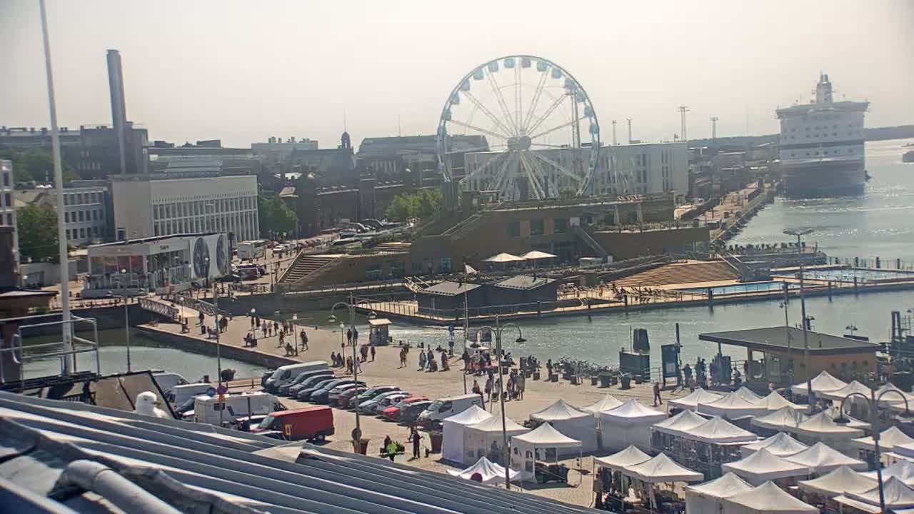 A harbor scene on a hazy day shows a Ferris wheel, a docked cruise ship, buildings, and numerous people walking along a waterfront promenade with market stalls set up.