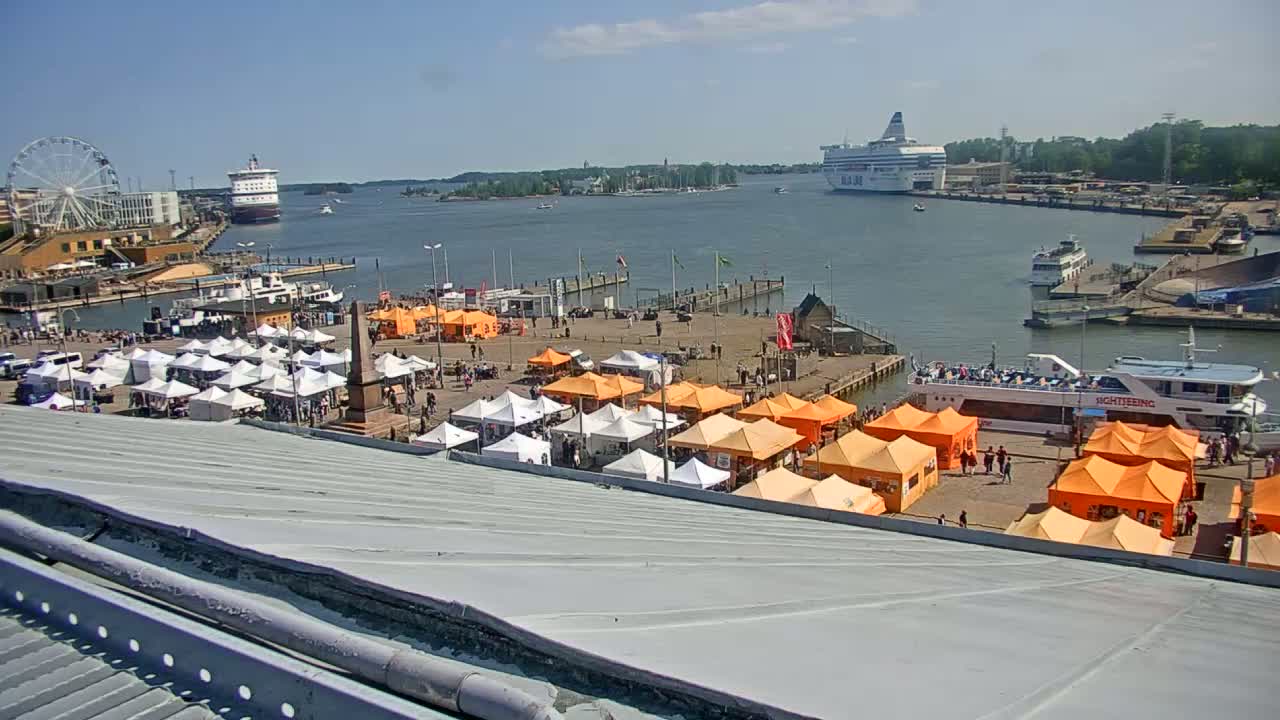 A harbor scene on a sunny day shows numerous white and orange market tents situated near a waterfront with several boats, ships, and a Ferris wheel in the background.