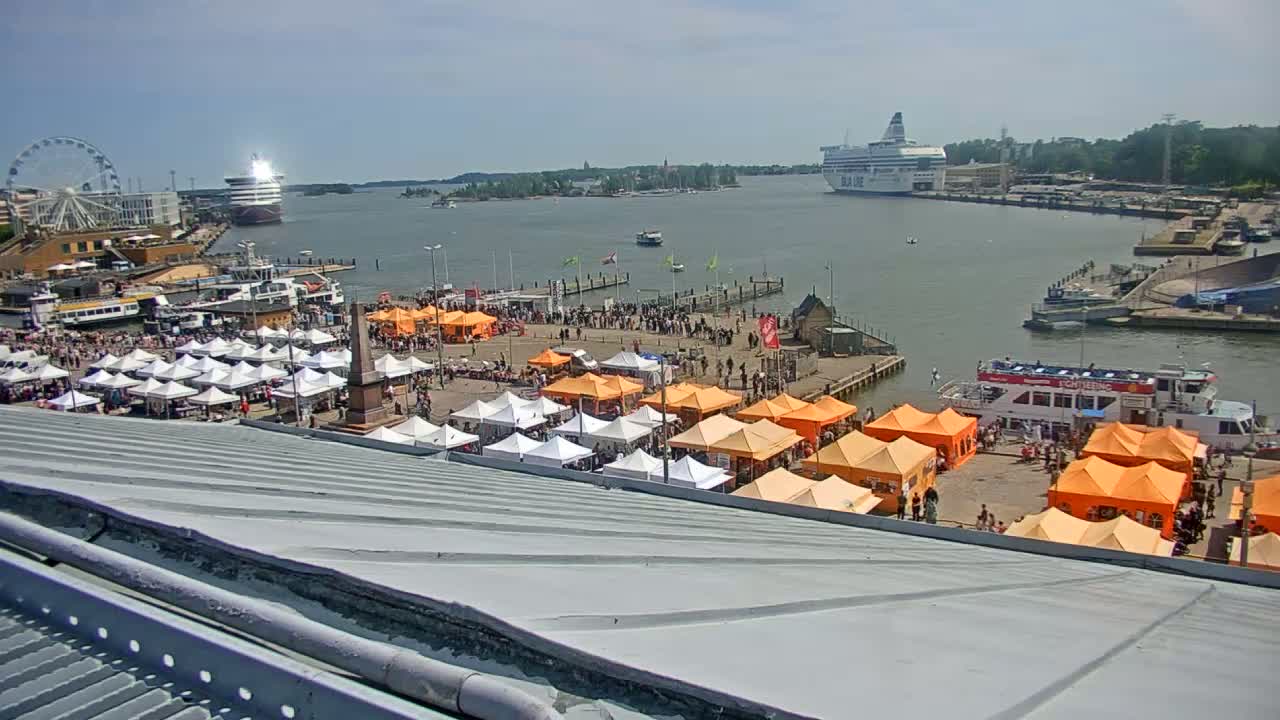 A harbor scene on a sunny day shows numerous market stalls, a Ferris wheel, several ships, and many people gathered near a waterfront.