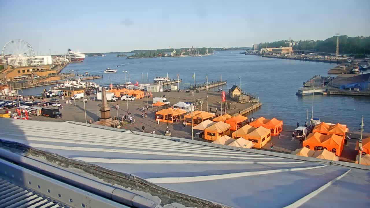 A harbor scene on a sunny day shows numerous orange tents set up in a plaza near a waterfront with boats, a Ferris wheel, and other buildings in the distance.