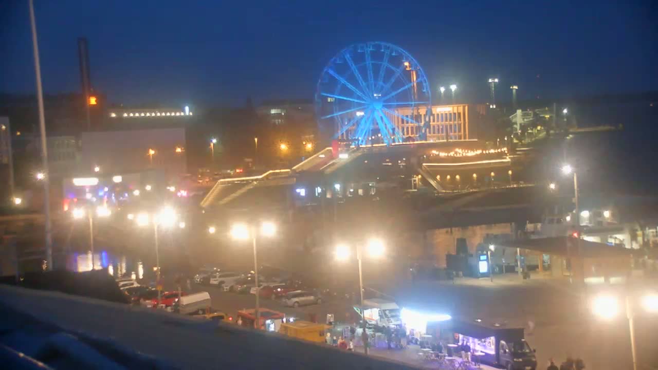 A harbor scene on a sunny day shows numerous orange tents set up in a plaza near a waterfront with boats, a Ferris wheel, and other buildings in the distance.