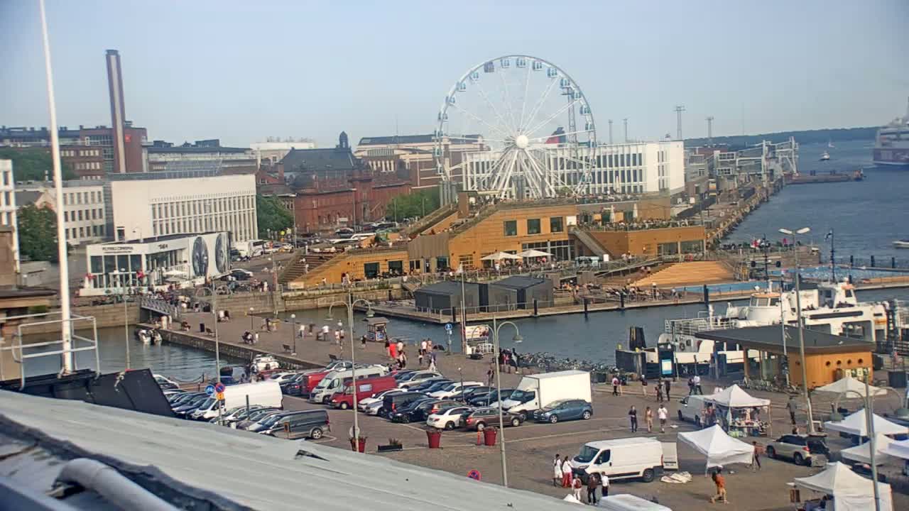 A waterfront cityscape on a sunny day features a Ferris wheel, buildings, parked cars, a ferry, and people walking along a harbor.