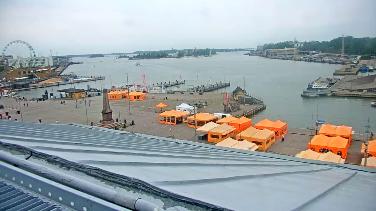 An overcast day reveals a harbor scene with numerous orange tents set up in a plaza next to a body of water, a Ferris wheel in the distance, and several boats docked.