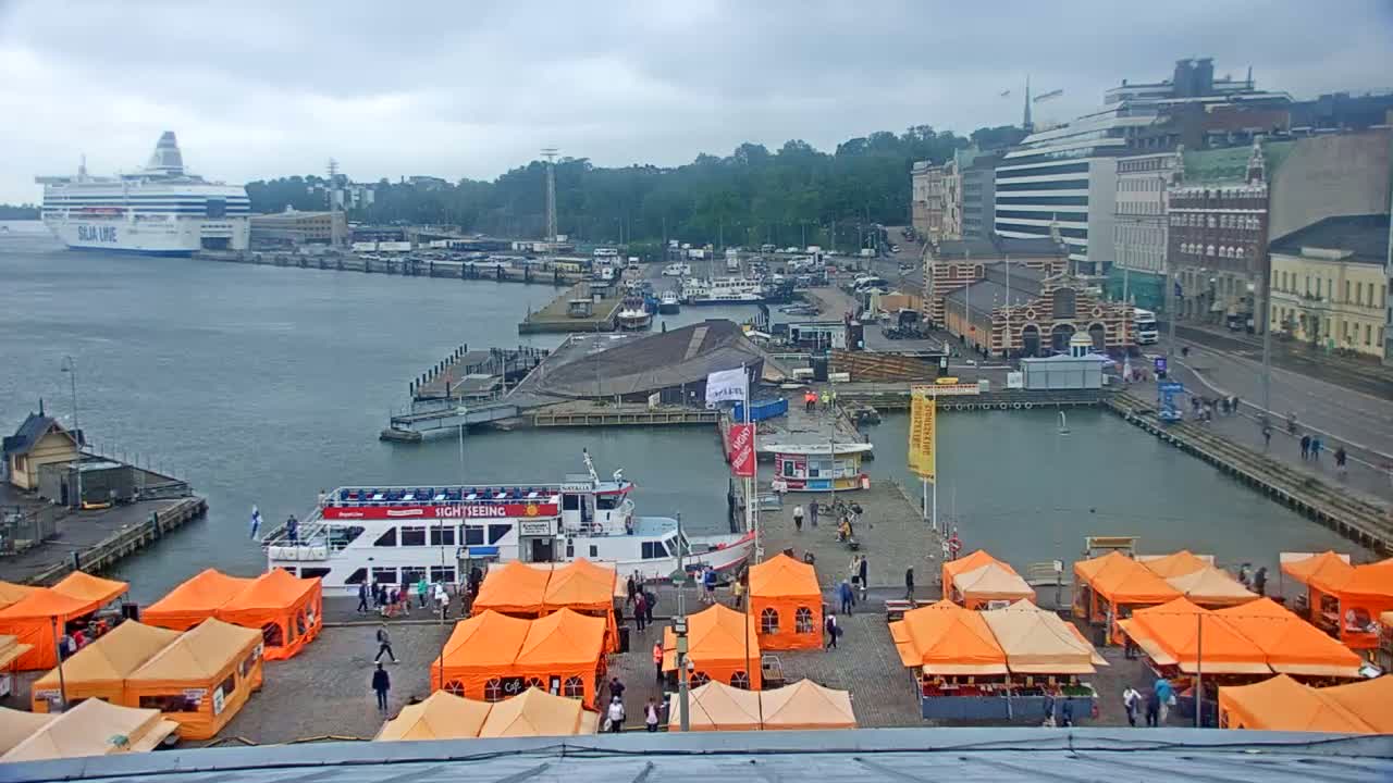 A harbor scene on an overcast day shows a large ferry, several smaller boats, orange market tents, and buildings along the waterfront.
