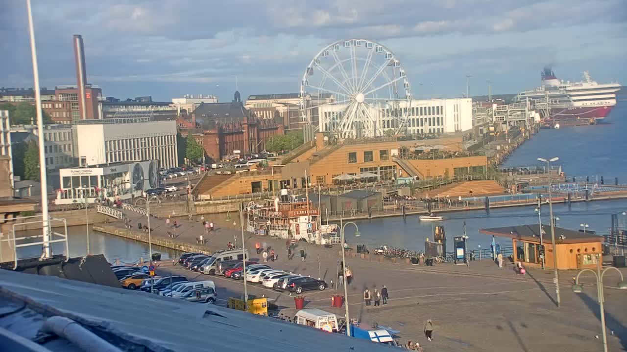 A harbor scene on a partly cloudy day shows a Ferris wheel, a docked cruise ship, buildings, and people enjoying the waterfront.