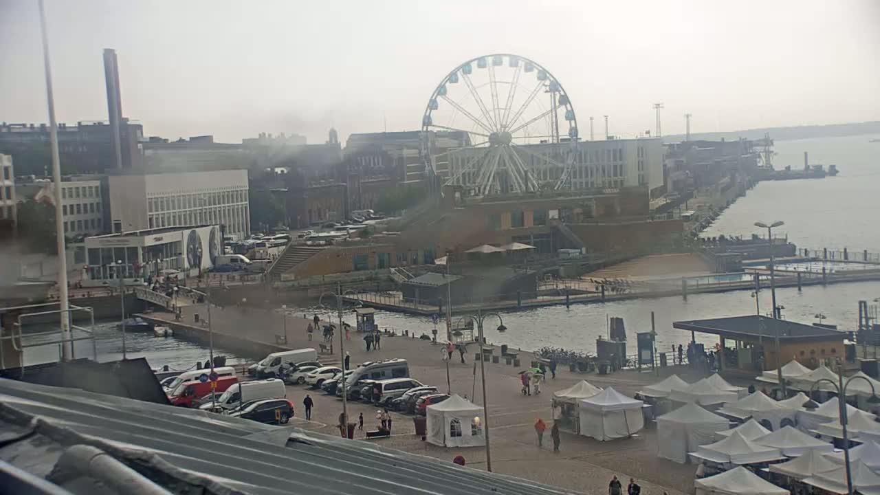 A waterfront scene on an overcast day shows a Ferris wheel, buildings, parked cars, people walking, and numerous white tents near a body of water.