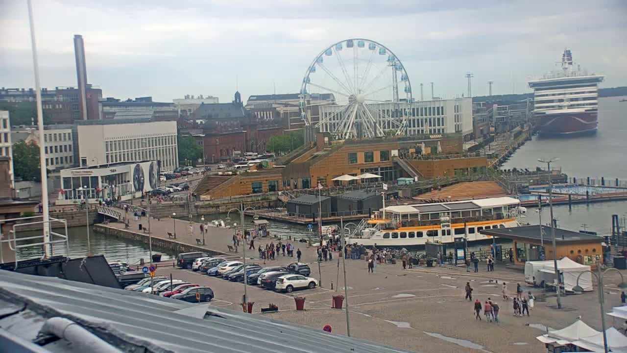 A harbor scene on an overcast day shows a Ferris wheel, a cruise ship, several buildings, a passenger boat, cars in a parking lot, and many people walking around.
