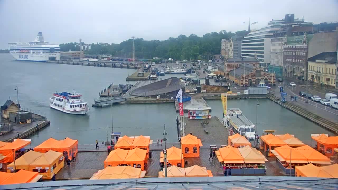 A harbor scene on an overcast day shows a large ferry, several smaller boats, buildings, and numerous orange market tents on a dock.