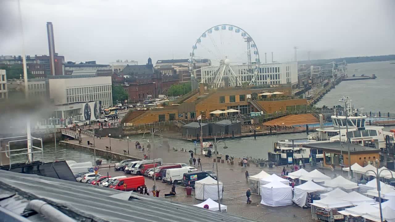 A waterfront city scene on a rainy day shows a Ferris wheel, buildings, boats, market stalls, and parked cars.