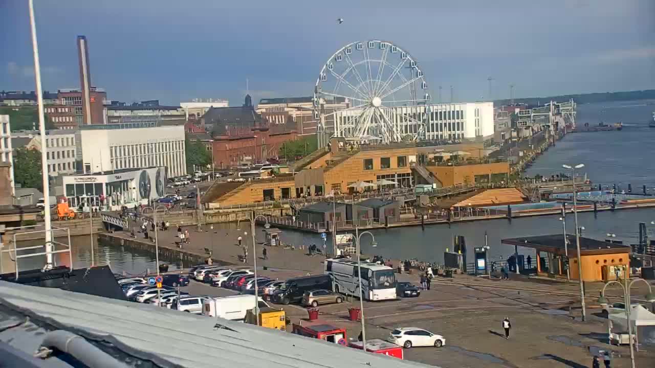 A waterfront scene on a partly sunny day shows a Ferris wheel, buildings, a harbor with boats, and a parking lot filled with cars and a bus.