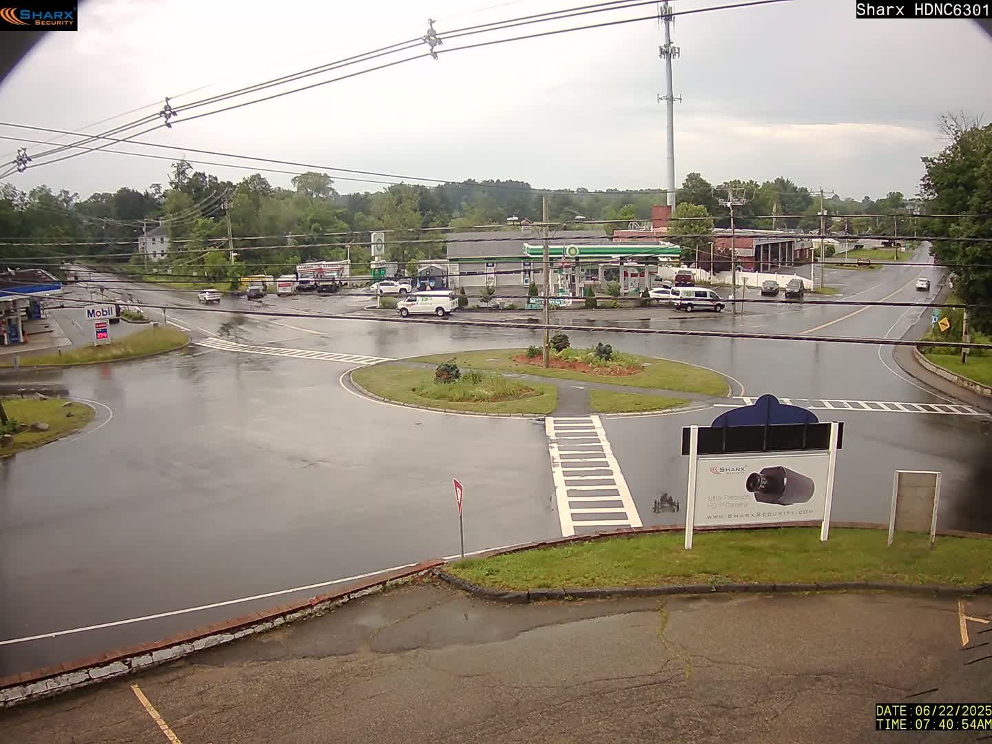 A wet roundabout and surrounding roads with several vehicles, gas stations, and buildings under an overcast sky.
