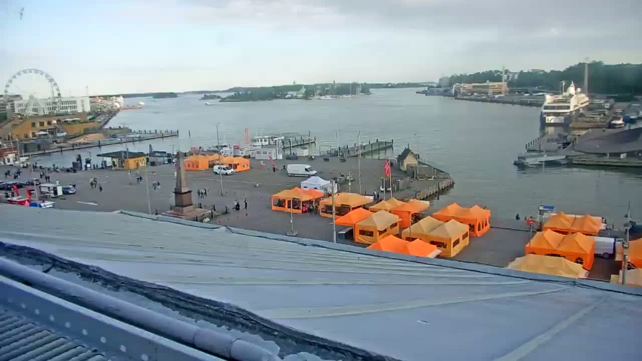 A harbor scene with numerous orange tents set up in a square, a Ferris wheel in the distance, and several boats docked, under an overcast sky.