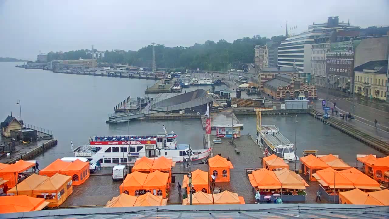 A harbor scene on a rainy day shows several boats, buildings, and orange market stalls along a waterfront.