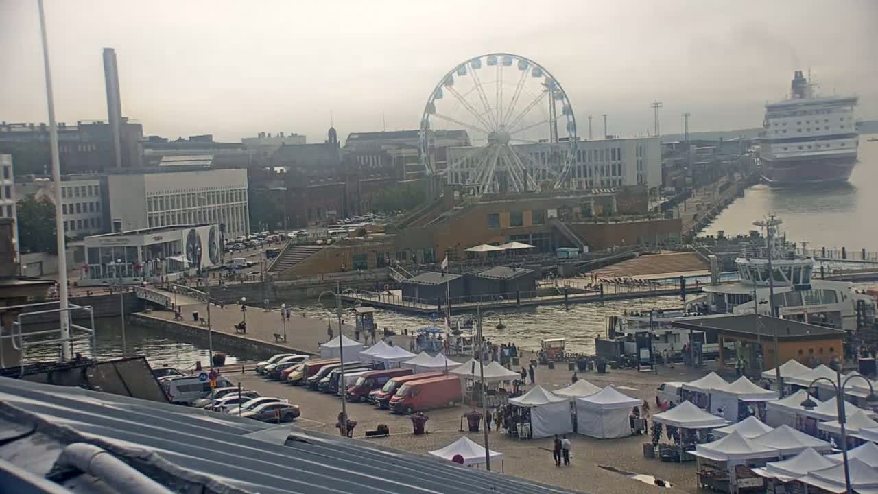 A Ferris wheel and a large cruise ship are visible in a harbor scene, on an overcast day, with numerous market stalls and parked cars.