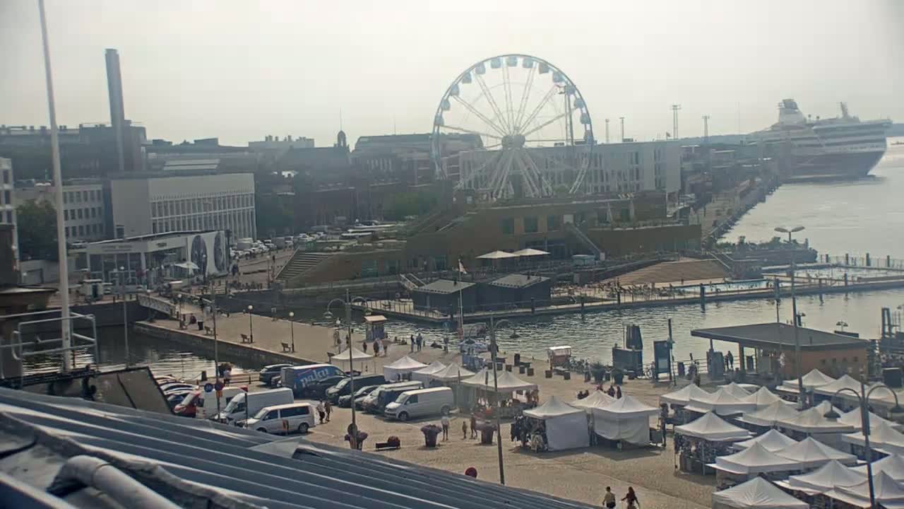 A waterfront area with a Ferris wheel, market stalls, parked cars, and a cruise ship in the distance on a hazy day.