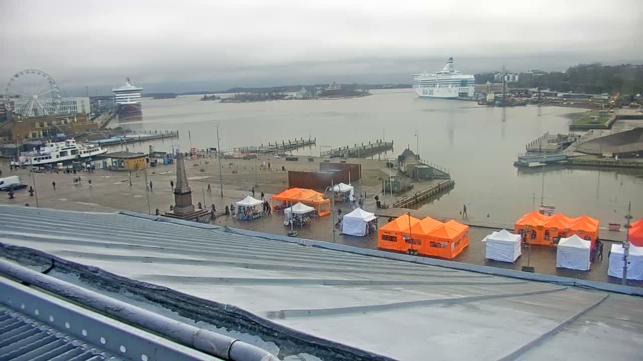 A bustling city harbor is seen from above on an overcast and wet day, featuring a market with many orange and white tents, pedestrians, several boats including a large white ferry, and a distant Ferris wheel under a cloudy sky.