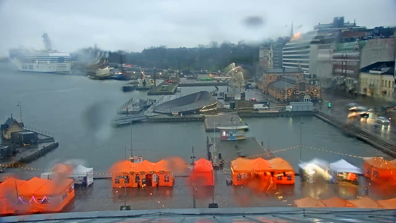Rainy weather blurs an overhead view of a bustling harbor featuring a large ferry, numerous boats, city buildings along the waterfront, and several orange and white market tents on the wet ground under an overcast sky.