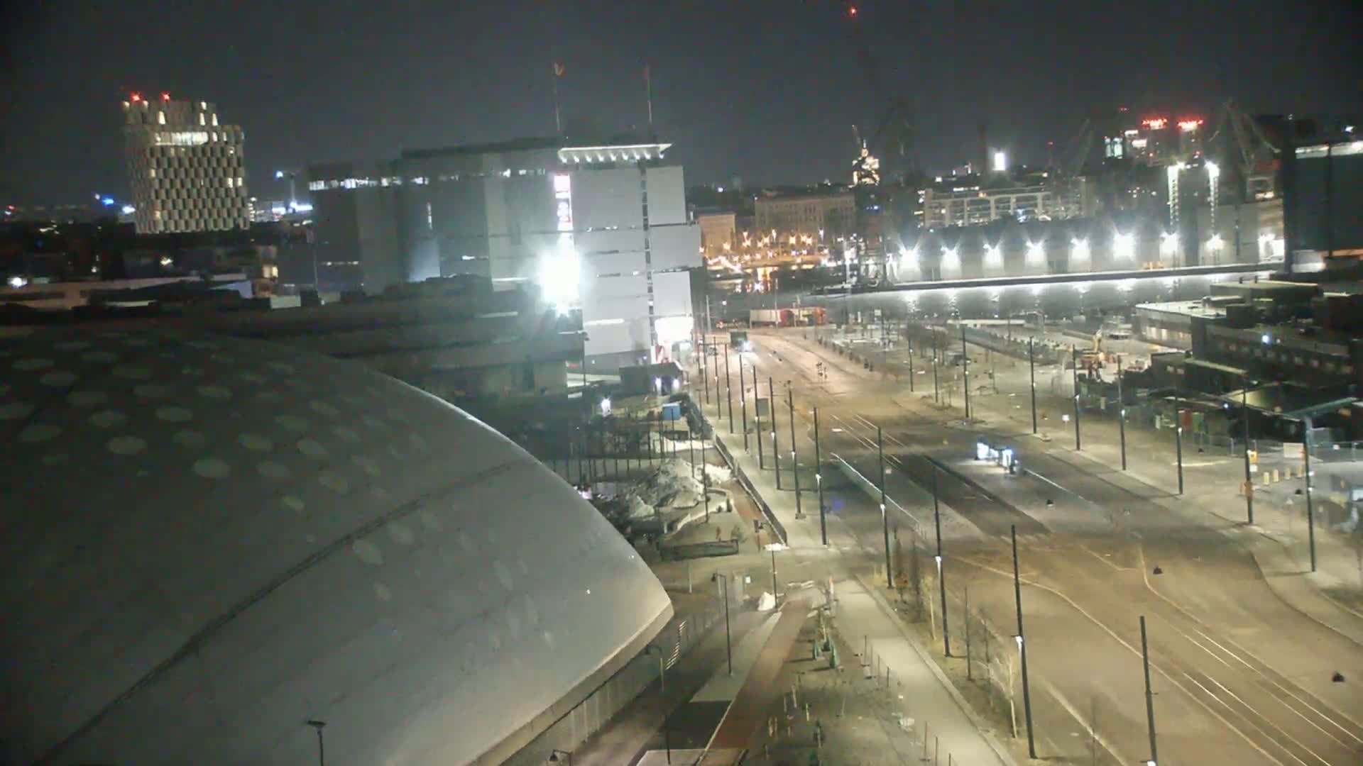 A nighttime cityscape showing a large domed building, several other buildings, and a wide, empty street with tram tracks, under clear skies.