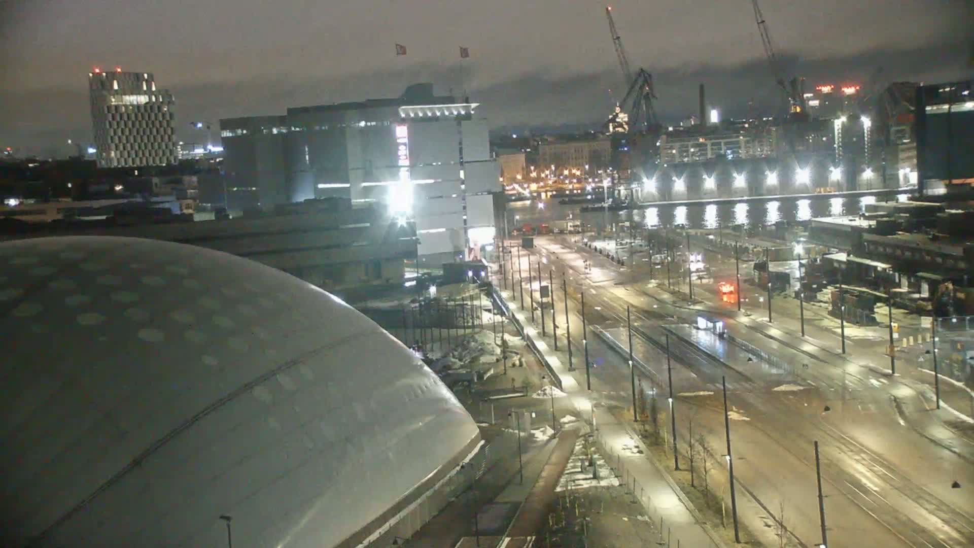 A nighttime, wet city scene shows a large domed structure next to a wide street with tram tracks, buildings, and cranes in the distance under a dark sky.