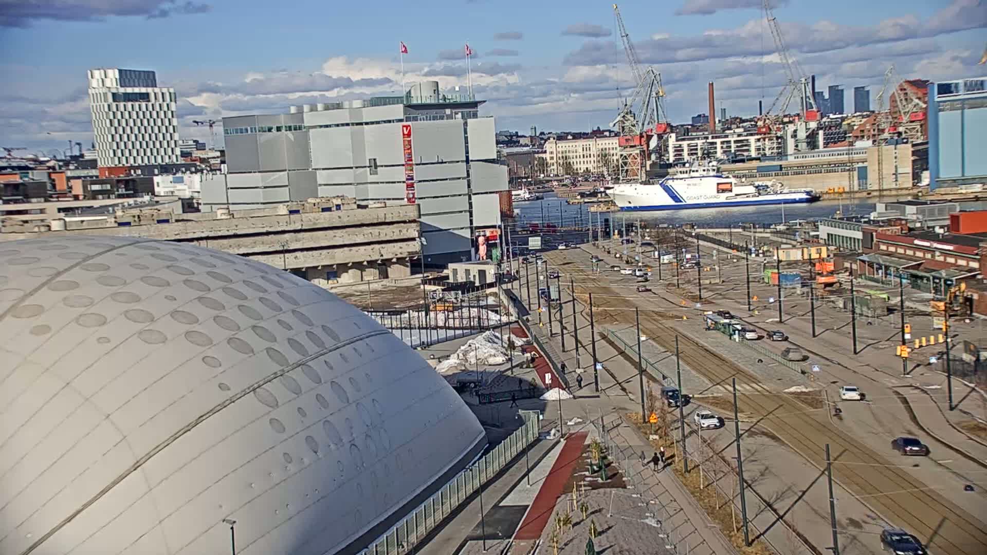 A large domed building is situated near a waterfront area with a coast guard ship, cranes, and other buildings under a partly cloudy sky.