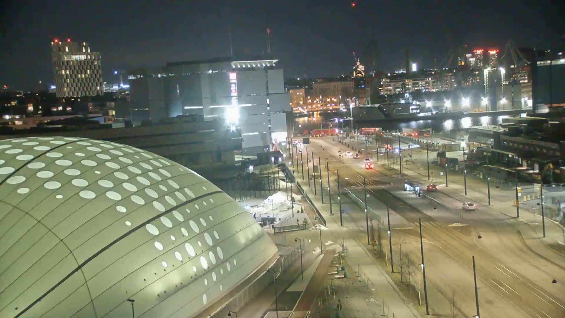 A nighttime aerial view of a city street with trams, sparsely populated with vehicles, leading to a waterfront area with industrial buildings and a domed structure with a pattern of white circles on its surface.